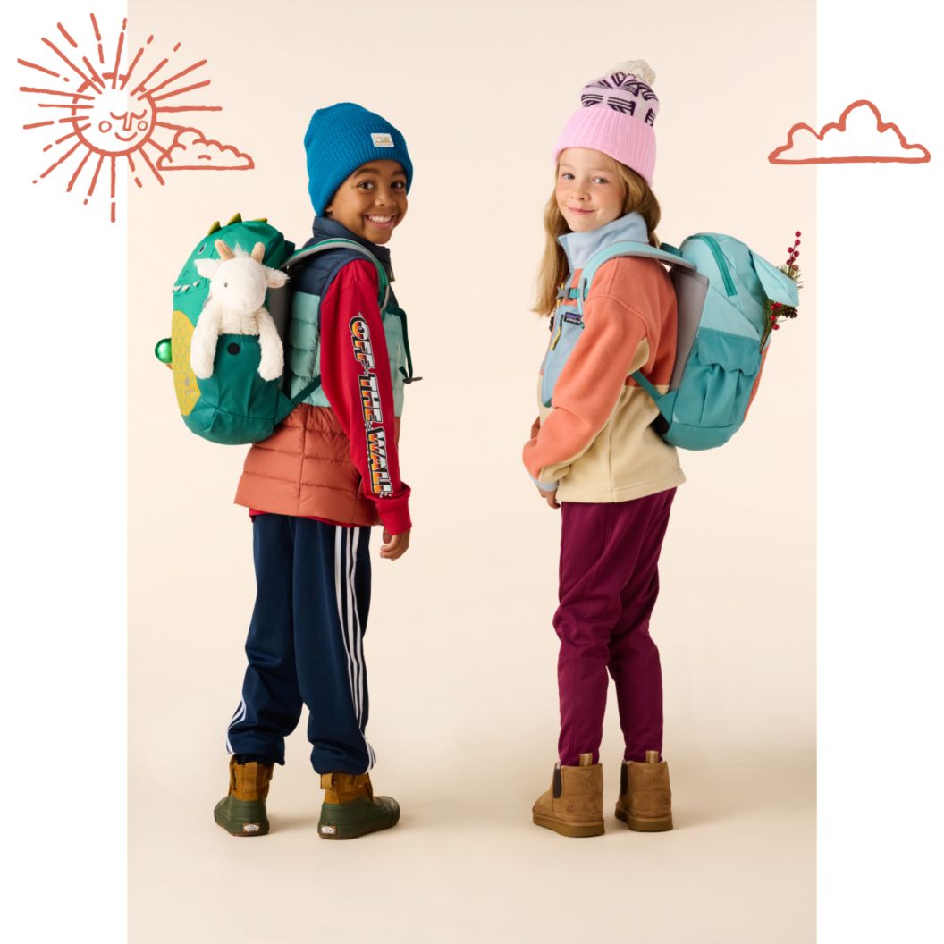 Two young kids wearing boots, fleece, beanies, and backpacks look over their shoulders with big smiles on their faces. 